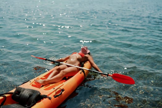 A woman is laying on a kayak in the water. The kayak is orange and has a paddle on it. The woman is wearing a red hat