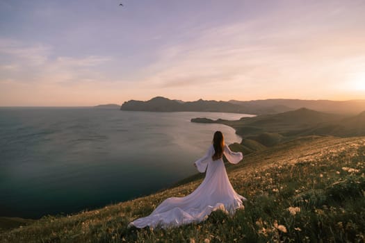 A woman in a white dress stands on a hill overlooking the ocean. The sky is a mix of blue and orange, creating a serene and romantic atmosphere