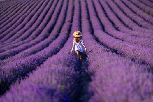 A woman is walking through a field of purple flowers. The field is vast and stretches out in front of her. The woman is wearing a blue dress and a straw hat