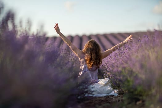 A woman is sitting in a field of lavender flowers. She is stretching her arms out in front of her