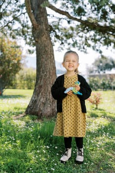 Little smiling girl with a soft toy in her hands stands under a tree on a green meadow. High quality photo