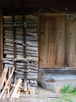 A rustic wooden cabin with a stack of firewood. There are chopped logs and a single shoe on the ground near the entrance.