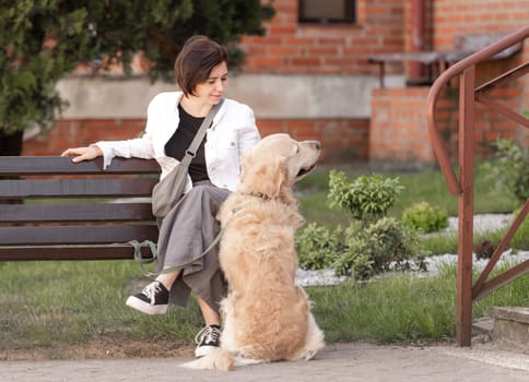 Young Woman Sits On Bench Near House With Golden Retriever In Spring