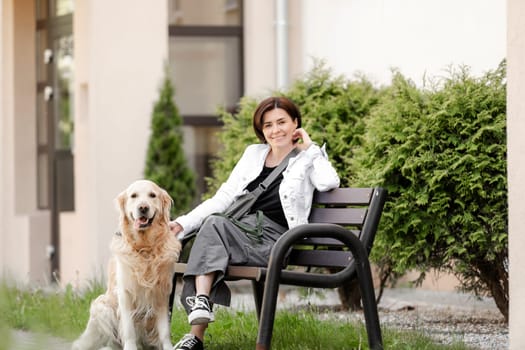 Young Woman Looks Into Camera While Sitting On Bench Near House With Golden Retriever In Spring