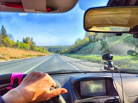 Car salon, windshield, hand on steering wheel and landscape in sunny day. View from driver on nature and Road. Single traveler. Partial focus. Blurred