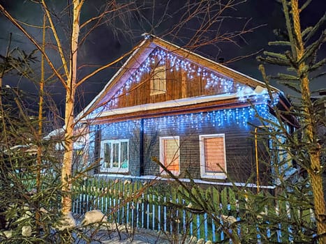 A charming wooden house adorned with blue and white Christmas lights glows amidst a snowy landscape, surrounded by trees