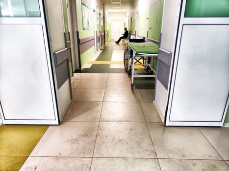 Sunlit Hospital Corridor With Gurney And Person Walking, Midday. A hospital corridor with a gurney, bright lighting, and a person walking in the background