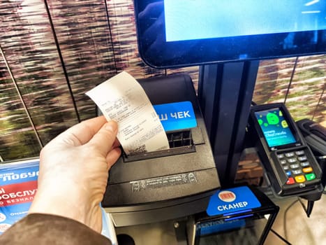 Kirov, Russia - April 17, 2024: Customer Holding Receipt at Checkout With Scales and Cash Register. Hand presents a receipt with scales and register