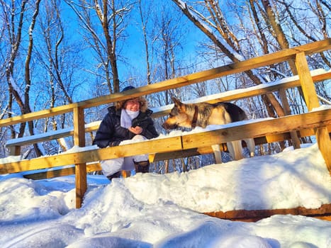 Adult girl or mature lady walking with shepherd dog, taking selfies in winter nature landscape. Middle aged woman and big shepherd dog on wooden bridge in cold day. Friendship, love, communication