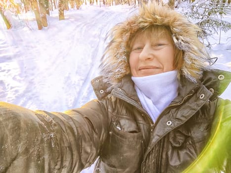 A cheerful middle aged woman in a winter coat with fur, scarf taking selfie outdoors on nature in a forest or park in beautiful sunny day