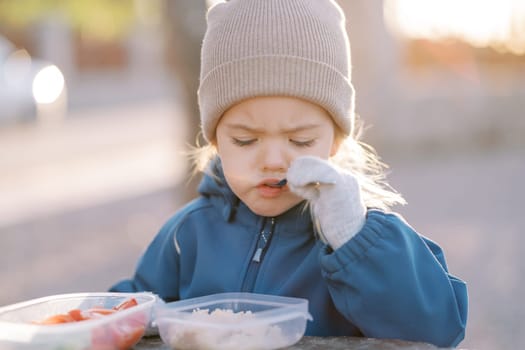 Little girl eats porridge with a spoon looking into her lunchbox while standing in the park. High quality photo