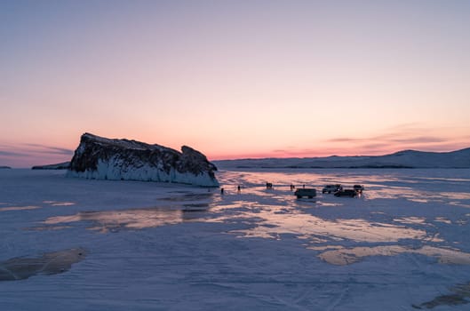 Aerial winter landscape of frozen lake Baikal. Groups of tourists got off the cars and walking around the rocky island in lake Baikal, walking on the crystal clear ice at sunrise. Famous tourist spot