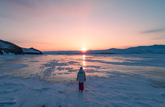 Aerial view on the young woman walking on the blue cracked ice of Baikal at beautiful orange sunrise. Sun reflections on the ice. Winter landscape of frozen Baikal.