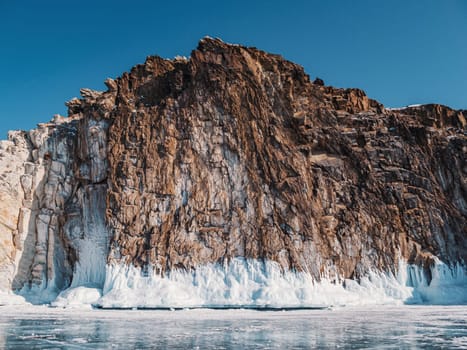 A majestic rocky cliff towers over the frozen surface of Lake Baikal, adorned with stunning ice formations and illuminated under a clear blue sky.