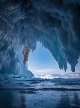 Inside a stunning ice cave on Lake Baikal, large icicles hang from the ceiling, creating a breathtaking winter landscape. Snow-covered mountains can be seen far in the distance.