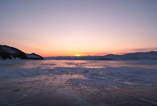 Snowy cracky ice of lake Baikal and the rocky island. Winter landscape of frozen Baikal at beautiful lilac sunrise. Sun reflections on the ice. Popular tourist spot. Tilt shot