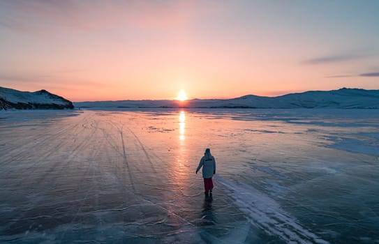 Aerial view on the young woman walking on the blue cracked ice of Baikal at beautiful orange sunrise. Sun reflections on the ice. Winter landscape of frozen Baikal.