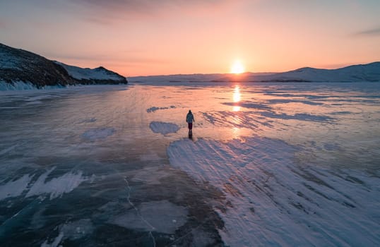 Aerial view on the young woman walking on the blue cracked ice of Baikal at beautiful orange sunrise. Sun reflections on the ice. Winter landscape of frozen Baikal.