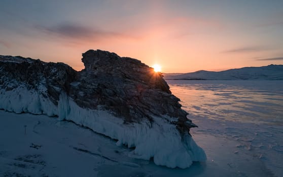 Aerial over the rocky island in lake Baikal. Winter landscape of frozen Baikal at beautiful orange sunrise. Sun reflections on the ice. Popular tourist spot.