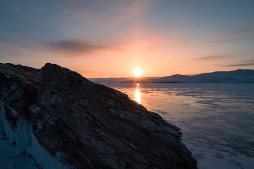 Aerial over the rocky island in lake Baikal. Winter landscape of frozen Baikal at beautiful orange sunrise. Sun reflections on the ice. Popular tourist spot.