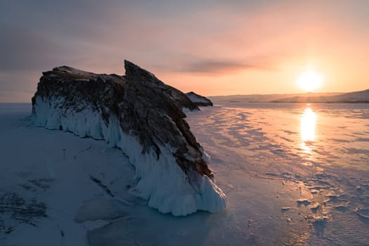 Aerial over the rocky island in lake Baikal. Winter landscape of frozen Baikal at beautiful orange sunrise. Sun reflections on the ice. Popular tourist spot.