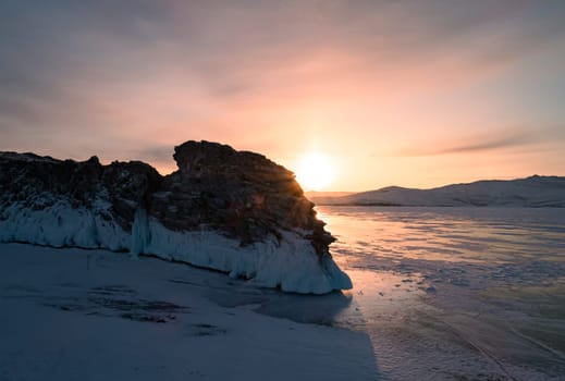 Aerial over the rocky island in lake Baikal. Winter landscape of frozen Baikal at beautiful orange sunrise. Sun reflections on the ice. Popular tourist spot.