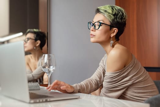 a beautiful girl with short hair and glasses is sitting indoors at a laptop, with her feet on the table, chatting and working online with a glass of water