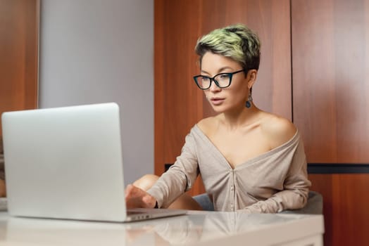 a beautiful girl with short hair and glasses is sitting indoors at a laptop, with her feet on the table, chatting and working online with a glass of water