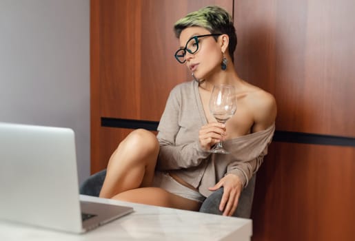 a beautiful girl with short hair and glasses is sitting indoors at a laptop, with her feet on the table, chatting and working online with a glass of water