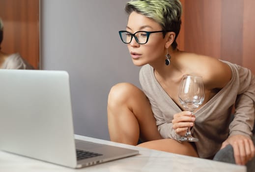 a beautiful girl with short hair and glasses is sitting indoors at a laptop, with her feet on the table, chatting and working online with a glass of water