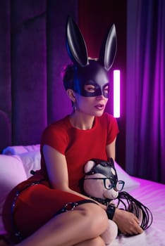 sexy girl in a bdsm rabbit mask and a bright red dress is sitting on a bed with a teddy bear in shoulder straps and leather belts in a neon light.