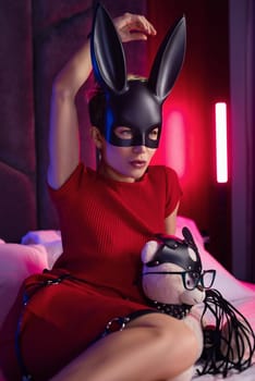 sexy girl in a bdsm rabbit mask and a bright red dress is sitting on a bed with a teddy bear in shoulder straps and leather belts in a neon light.