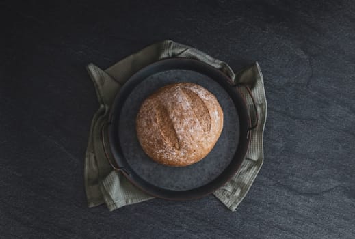 One round rye bread in a vintage metal plate with a kitchen napkin lies in the center on a black stone background, flat lay close-up. Bread baking concept.