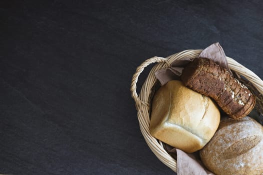 Three different varieties and shapes in an oval wicker basket with handles lie to the right on a black stone table with copy space on the left, flat lay close-up. The concept of baking bread.