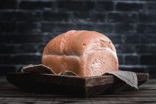 One square white bread in a wooden dish with a kitchen napkin lies on a brown wooden table against a blurred black brick wall, close-up view from below. The concept of baking bread.