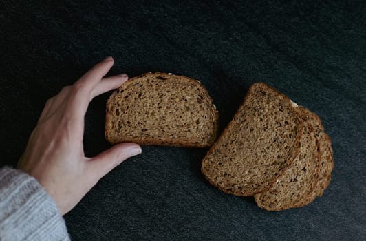 The hand of a young caucasian girl holds one slice of rye bread with sunflower seeds next to three slices on a black stone table, close-up side view. The concept of baking bread.