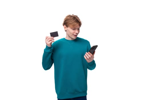 young handsome man with red hair dressed in a blue jacket holds a credit card and a phone.