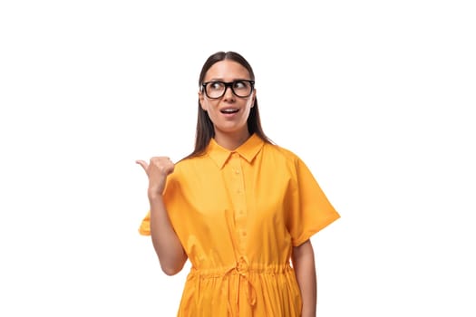 Caucasian young slender woman with black straight hair with glasses and in a yellow dress gesticulates with her fingers.