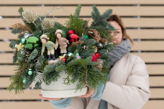 A woman is standing, holding a potted plant that is adorned with Christmas decorations. She appears to be showcasing the festive plant, which features sparkly baubles, ribbons, and lights.