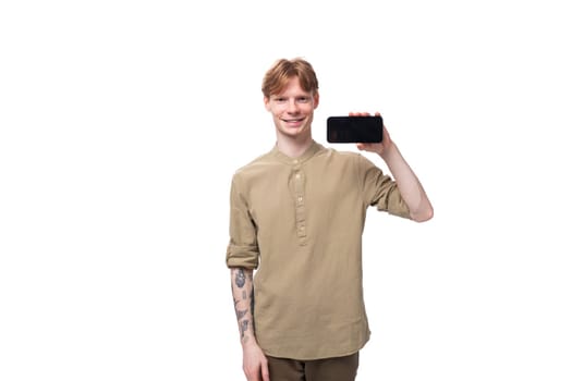 a young european guy with red hair is dressed in a fashionable beige shirt and brown trousers shows a horizontal smartphone.