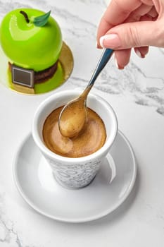 Female hand stirring rich crema atop freshly brewed espresso served with glossy green apple-shaped pastry on marble top. Moment of delightful indulgence of coffee and sweets
