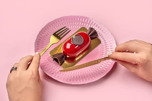 Woman preparing to enjoy artisanal candy-shaped mousse dessert with glossy red glaze served on textured pink plate, holding gold cutlery in hands. Cropped image