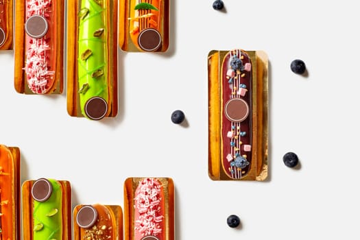 Assortment of tempting decorated French eclairs with vibrant colorful glazes topped with fresh berries, dried fruits, nuts and meringue pieces, arranged on golden serving cardboards on white, top view