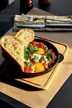 Appetizing savory shakshuka with sunny-side up eggs, spicy tomato sauce, hummus and fresh herbs, served with crusty toasts in cast iron pan on wooden board