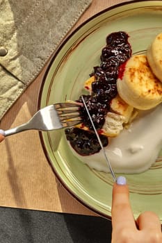 Close-up of female hands cutting into warm quark pancakes served with berry sauce and sour cream on decorative plate. Traditional Slavic sweet treat