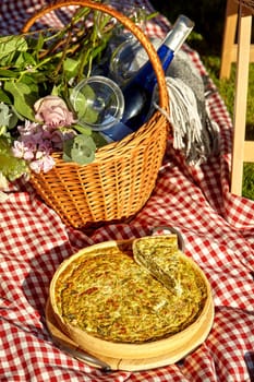 Appetizing savory quiche with cheese and spinach served on wooden board on red and white checkered blanket with wine bottle and glasses in wicker basket. Cozy romantic summer picnic on green grass