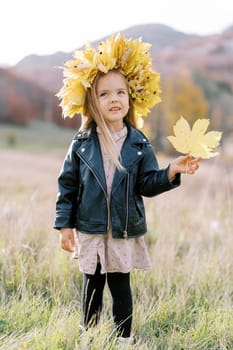 Little girl wearing a wreath of autumn leaves with a yellow maple leaf in her hand stands on the lawn. High quality photo