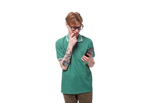 young smart caucasian red-haired man dressed in a green t-shirt carefully looks into a smartphone.