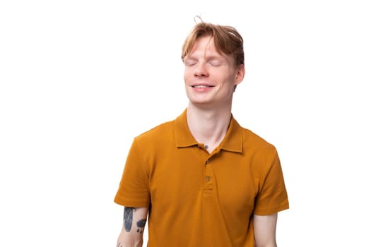 a young red-haired guy in an orange t-shirt shyly closed his eyes on a white background with copy space.