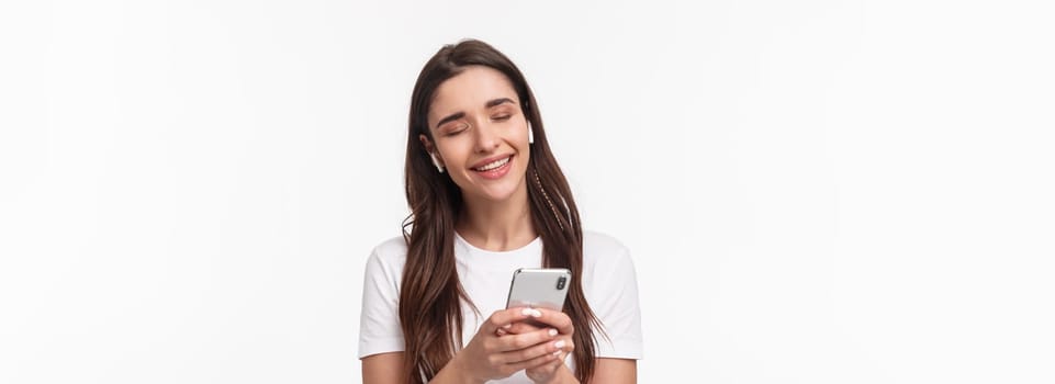 Close-up portrait of tender cute young brunette woman close eyes, shaking hand in rhythm music, smiling from delight, listening song in wireless earphones, holding smartphone.